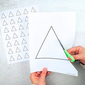 May include: Two sheets of white paper with black triangle outlines. One sheet has multiple small triangles, the other has a large triangle being cut with green-handled scissors. The image is on a light grey surface.