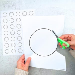 Könnte beinhalten: Eine Person schneidet mit einer grün-griffigen Schere einen großen Kreis aus einem weißen Blatt Papier. Ein weiteres Blatt Papier mit mehreren schwarz umrandeten Kreisen ist sichtbar. Das Bild ist vor einem hellgrauen Hintergrund.