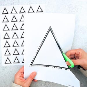 Op de afbeelding: Een persoon knipt met een groene schaar een grote driehoek uit een wit vel papier. De driehoek heeft een zwarte omtrek met een stippellijn. Een ander vel papier met meerdere zwarte driehoeken is zichtbaar.