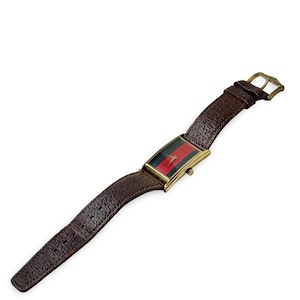 Puede incluir: Un reloj de pulsera rectangular dorado con una correa de cuero marrón. La esfera del reloj presenta un diseño de rayas rojas y verdes. La correa es de cuero marrón oscuro texturizado con una hebilla dorada.