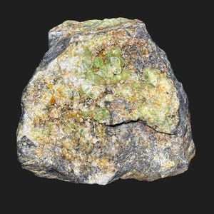 Puede incluir: Un espécimen de roca en bruto con un exterior gris oscuro y marrón. Contiene grupos de formaciones cristalinas de color verde claro incrustadas en la roca. El espécimen tiene una apariencia natural y terrosa, con variaciones en textura y color.