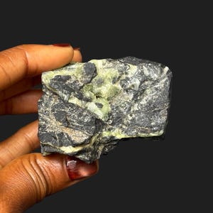 Puede incluir: Una formación rocosa tosca con una mezcla de estructuras cristalinas gris oscuro y verde claro. La roca se sostiene sobre un fondo oscuro, mostrando sus texturas naturales y variaciones de color. La apariencia general sugiere un espécimen mineral en bruto y sin pulir.