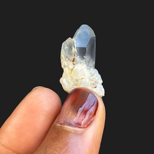 Puede incluir: Un grupo de cristales de cuarzo transparente con una estructura puntiaguda y facetada. El cristal se sostiene sobre un fondo negro, mostrando su forma natural y claridad. El cristal mide aproximadamente 2,5 cm de altura.