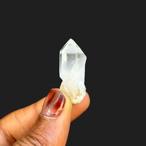 Puede incluir: Un cristal de cuarzo transparente con una punta y una base rugosa de color blanquecino. El cristal se sostiene sobre un fondo negro, mostrando su cualidad translúcida y su forma natural. El cristal mide aproximadamente 3,8 cm de largo.