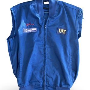 May include: A blue sleeveless jacket with a zipper closure. The jacket features the name "John" embroidered in red script, along with patches that read "Beverage Distributors" and "Lite Beer". The jacket has a ribbed collar and hem.