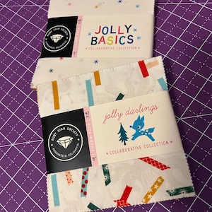 May include: Two fabric charm packs, "Jolly Basics" and "Jolly Darlings", each containing 42 pieces. The "Jolly Basics" pack has a white background with colorful stars. The "Jolly Darlings" pack features a white background with colorful ribbon designs and a deer graphic.