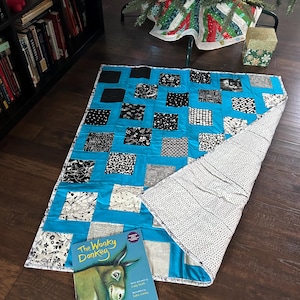 May include: A blue and white patchwork quilt with black squares. The quilt is folded in half and a children's book titled "The Wonky Donkey" is lying on top of it.