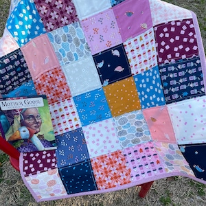 May include: A patchwork quilt with a pink border and a variety of colorful patterns, including dice, houses, and geometric shapes. The quilt is sitting on a red chair with a book titled "Mother Goose Nursery Rhymes" visible in the foreground.