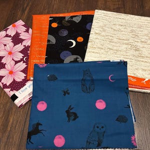 May include: A collection of fabric pieces with various patterns. One fabric features large pink flowers on a dark purple background. Other fabrics display celestial, woodland, and abstract designs in orange, black, blue, and beige.