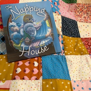 May include: A book titled "The Napping House" by Audrey Wood and Don Wood. The book cover features a blue and white illustration of a house with a family of animals sleeping inside. The book is lying on a colorful patchwork quilt.