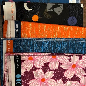 May include: A stack of fabric pieces with various patterns. Designs include a blue fabric with a silhouette, a black fabric with a moon and stars, an orange fabric with gold streaks, a blue textured fabric, and a floral pattern.