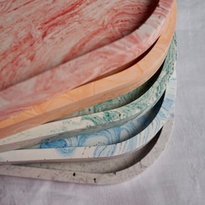 Peut inclure: Une pile de plateaux décoratifs aux couleurs variées, dont rose, pêche, vert, bleu et gris. Chaque plateau a une forme rectangulaire arrondie avec un motif marbré. Les plateaux sont empilés, mettant en valeur leurs différents motifs.
