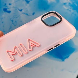 May include: A pink phone case with a clear back and a black camera ring. The case has the name "MIA" printed on the back in a pink outline.