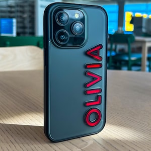 Peut inclure: Une coque de téléphone noire avec un dos transparent et un contour rouge du nom "Olivia" au dos.
