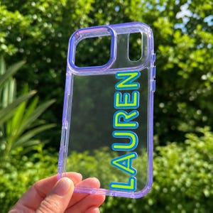 May include: A clear phone case with a purple frame and the name LAUREN printed vertically in bright lime green letters with a blue outline. The case features a rectangular camera cutout and a smaller vertical cutout for the flash and sensors.