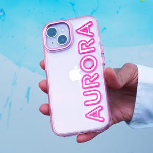 May include: Clear phone case with a pink border and the name "AURORA" in pink letters. The case is held against a blue and white background. The phone has a silver Apple logo.