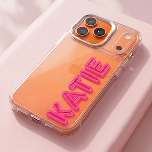 May include: Clear phone case with an orange interior and the name "KATIE" in bright pink, three-dimensional letters. The case has a clear border and a camera module with three lenses.
