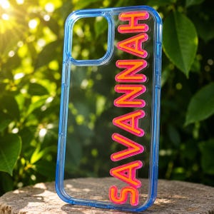 Op de afbeelding: Een transparant telefoonhoesje met een blauwe rand, voorzien van de naam SAVANNAH in verhoogde, felroze en oranje blokletters die verticaal langs de rechterkant van het rechthoekige frame voor een mobiel apparaat zijn geplaatst.