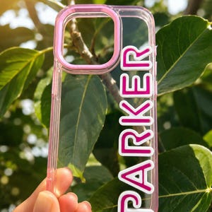 May include: A clear phone case with a pink camera lens border and the name PARKER written vertically in bold, raised pink letters with a white outline. The case features a transparent back and sides, showcasing a rigid, protective design.