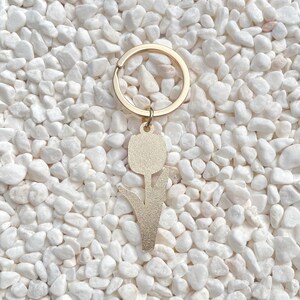 May include: A gold-colored metal keychain featuring a tulip design. The keychain has a round ring and a pendant shaped like a tulip flower. The background is composed of small, white pebbles.