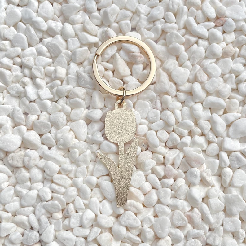 May include: A gold-colored metal keychain featuring a tulip design. The keychain has a round ring and a pendant shaped like a tulip flower. The background is composed of small, white pebbles.