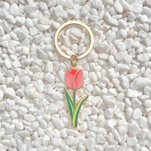 May include: A gold-colored metal keychain with a pink tulip charm. The tulip has a red top and green stem and leaves. The keychain is set against a background of white pebbles.