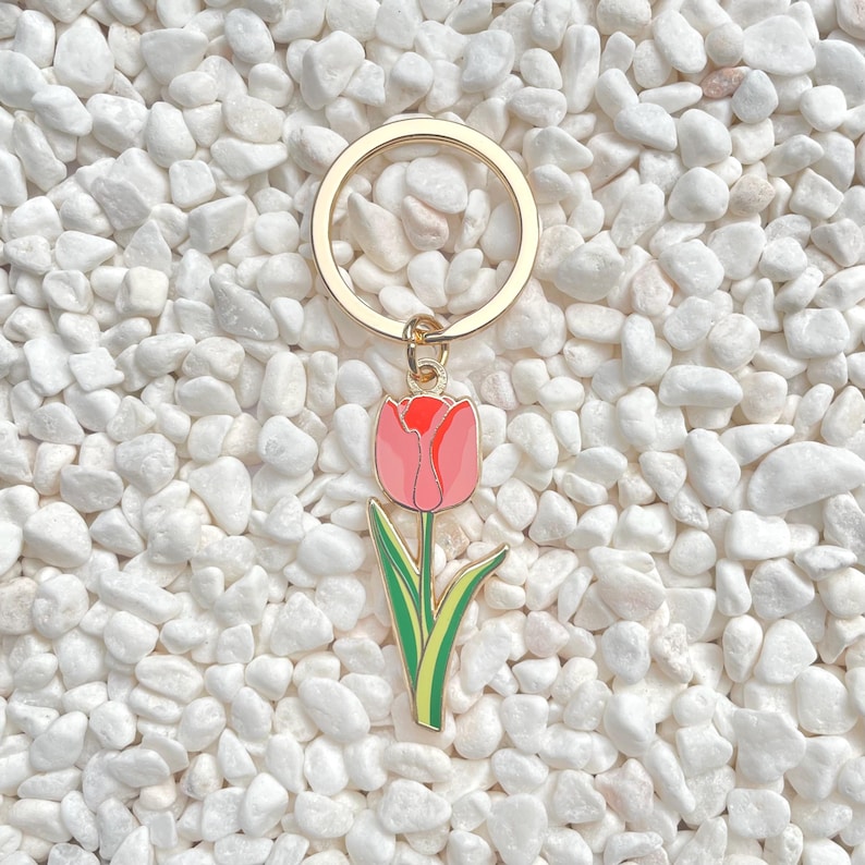 May include: A gold-colored metal keychain with a pink tulip charm. The tulip has a red top and green stem and leaves. The keychain is set against a background of white pebbles.