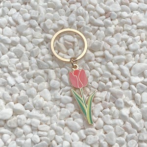 May include: A gold-colored metal keychain with a pink tulip charm. The tulip charm has green leaves and a pink flower. The keychain is set against a background of white pebbles.