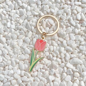 May include: A gold-colored metal keychain with a pink tulip charm. The tulip charm has a red top, pink petals, green leaves, and a yellow outline. The keychain is set against a background of white pebbles.