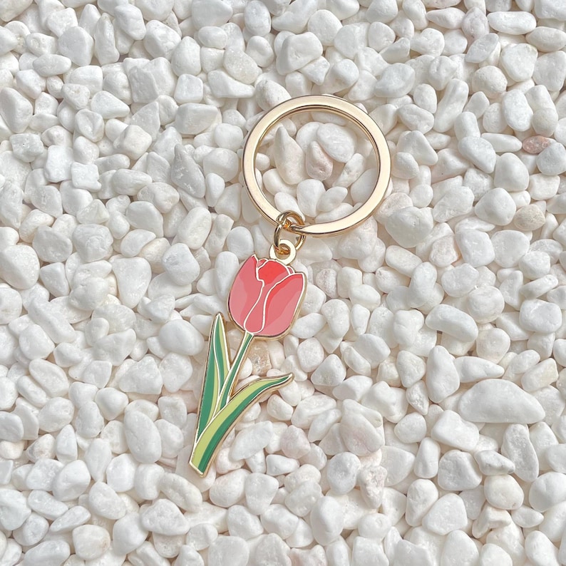 May include: A gold-colored metal keychain with a pink tulip charm. The tulip charm has a red top, pink petals, green leaves, and a yellow outline. The keychain is set against a background of white pebbles.