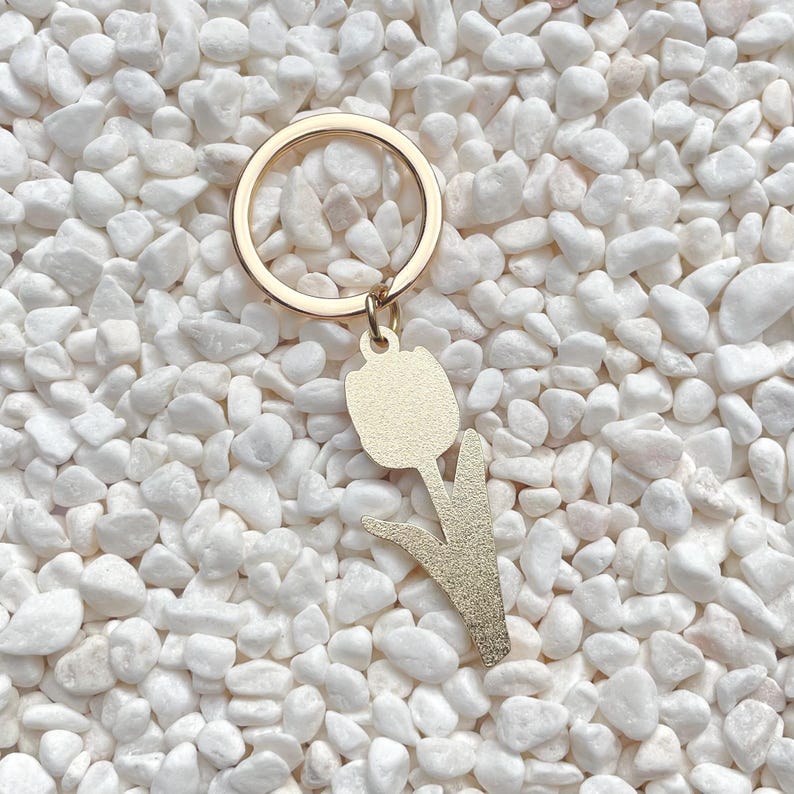 May include: Gold-colored metal keychain featuring a tulip design. The keychain has a round ring and a pendant shaped like a tulip flower with a stem and leaves. The background is white pebbles.
