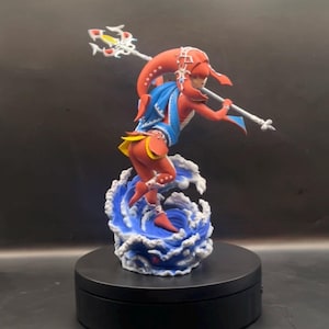 May include: A figurine of a red-skinned humanoid character with a long headpiece, wearing a blue cape and yellow accents, holding a silver trident. The character is posed dynamically atop a sculpted base of swirling blue and white water waves.
