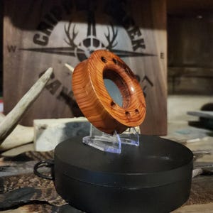 May include: A wooden, donut-shaped object with a polished, reddish-brown finish. The object has a central hole and several small, evenly spaced holes around its perimeter. It is displayed on a clear stand against a blurred background.