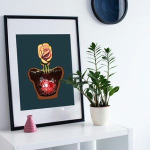 May include: Framed art print featuring a digital illustration of a rose growing in a pot. The rose has yellow and red petals. The pot has a glowing red center. A small pink vase sits below the artwork.