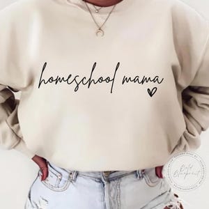 May include: Cream-colored sweatshirt with the words "homeschool mama" in a cursive font, accompanied by a small heart. The sweatshirt is a neutral color, and the text is in black. The design is simple and elegant.