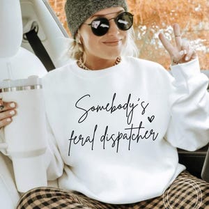 May include: White sweatshirt with black text that reads "Somebody's feral dispatcher" with a small heart symbol.