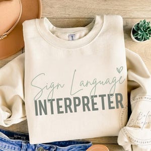 Può includere: Felpa color crema con le parole "Sign Language Interpreter" in verde scritto e lettere maiuscole. Un piccolo cuore verde è sopra la parola "Interpreter". Un cappello color cammello e jeans sono visibili sullo sfondo.