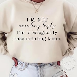 May include: A white sweatshirt with the text "I'M NOT avoiding tasks I'm strategically rescheduling them" printed on the front.