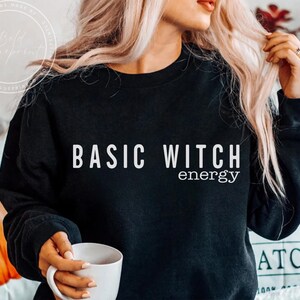 May include: Black sweatshirt with the text "BASIC WITCH energy" in white. The sweatshirt is a crew neck style. A white mug is held in the hand. The design is a fun, casual style.