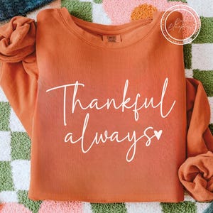 May include: An orange Comfort Colors sweatshirt with the words "Thankful always" in white script. A small white heart is next to the word "always". The sweatshirt is folded and placed on a patterned surface.