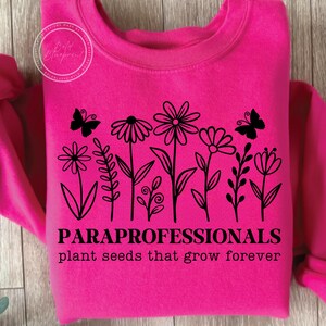 May include: Hot pink sweatshirt with a black floral design and two butterflies. The text "PARAPROFESSIONALS plant seeds that grow forever" is printed below the flowers. The sweatshirt is made of a soft material.