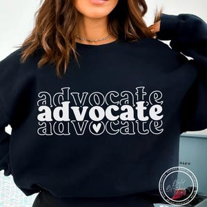 May include: A black sweatshirt featuring the word "advocate" in white, repeated three times. The middle "advocate" has a heart symbol instead of the letter "o". A comfortable, casual style.
