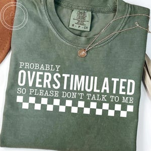 May include: Olive green t-shirt with the text "PROBABLY OVERSTIMULATED SO PLEASE DON'T TALK TO ME" in white. The shirt has a checkered pattern at the bottom and a Comfort Colors tag. A gold necklace with a pendant is draped across the shirt.