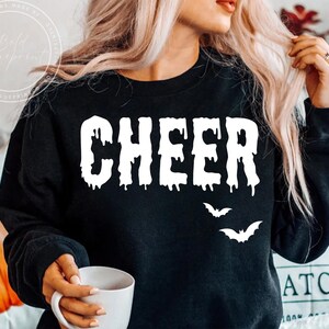 Op de afbeelding: Zwarte sweatshirt met het woord "CHEER" in druipende witte letters en twee witte vleermuis silhouetten. Halloween thema.
