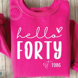 Peut inclure: Un sweat-shirt rose vif avec l'inscription blanche "hello FORTY", un cœur au-dessus du "o" de hello. L'année "est. 1986" est imprimée en dessous. Un vêtement décontracté pour un anniversaire.