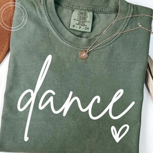 May include: An olive green t-shirt with the word "dance" in white script and a small white heart. A gold-coloured necklace with a pendant rests on the shirt. The shirt has a Comfort Colors label.