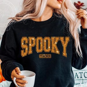 May include: Black sweatshirt with the word "SPOOKY" in gold glitter lettering. Below the word "SPOOKY" is the word "TEACHER" in a smaller gold glitter font. The sweatshirt is a crew neck style.