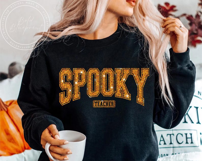May include: Black sweatshirt with the word "SPOOKY" in gold glitter lettering. Below the word "SPOOKY" is the word "TEACHER" in a smaller gold glitter font. The sweatshirt is a crew neck style.