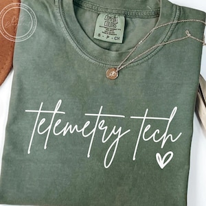 Op de afbeelding: Olijfgroen t-shirt met de woorden "Telemetry Tech" in witte letters en een klein hartje. Een gouden ketting met een hanger ligt op het shirt. Het shirt heeft een Comfort Colors label.