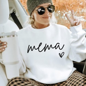 May include: White sweatshirt with the word "mema" in black script and a small heart. The sweatshirt is worn with a gray beanie, sunglasses, and a gold chain. The person is holding a white tumbler.